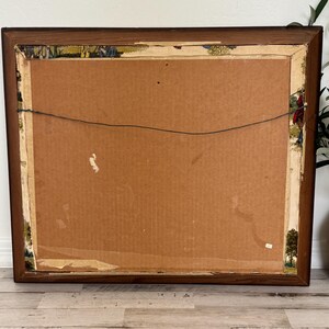 May include: Back of a framed artwork with a brown wooden frame. The artwork's backing is cardboard, with a wire for hanging. The edges of the artwork show remnants of a decorative border with a landscape design.