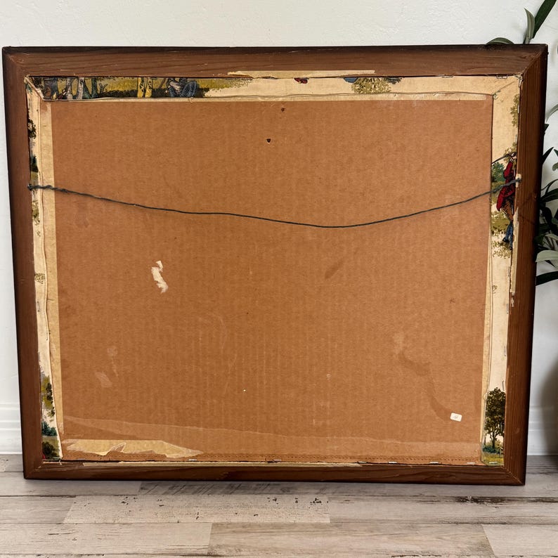 May include: Back of a framed artwork with a brown wooden frame. The artwork's backing is cardboard, with a wire for hanging. The edges of the artwork show remnants of a decorative border with a landscape design.