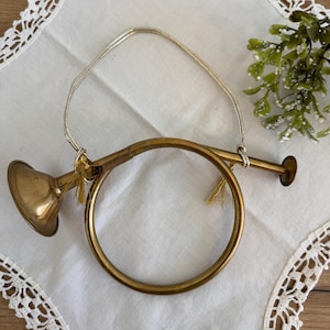 May include: A vintage brass bugle ornament with a gold cord for hanging. The bugle has a bell-shaped opening and a circular body. Small gold tassels are attached to the cord. The ornament is set on a white lace-trimmed cloth.