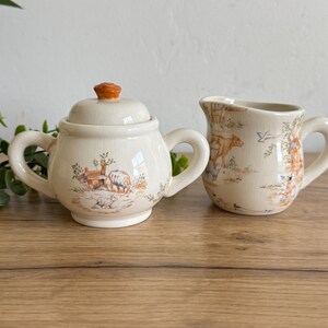 May include: A cream-colored ceramic sugar bowl with a lid and a matching creamer. Both pieces feature a pastoral scene with sheep and trees in shades of brown and orange. The sugar bowl has two handles.