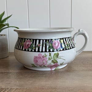 May include: A white ceramic chamber pot featuring a black and white vertical striped band around the upper section, decorated with pink rose floral patterns and green leaves. The vessel includes a curved handle on the right side and a wide rim.