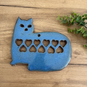 May include: A blue ceramic cat-shaped trivet with heart cutouts. The trivet has a glossy finish and a small hole at the top for hanging. The cat's body features eight heart-shaped cutouts. The trivet is placed on a wooden surface.