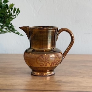 May include: A brown ceramic pitcher with a handle and spout. The pitcher has a glossy finish and a decorative floral pattern around the base. The pitcher is sitting on a wooden surface.