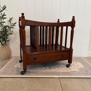 May include: A dark brown wooden magazine rack featuring a slatted design with vertical spindles, four corner posts with finials, and a single bottom drawer equipped with two circular metal pulls. The unit is supported by four small black caster wheels.