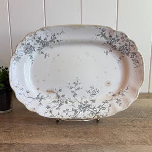 May include: An oval ceramic serving platter with a scalloped edge, featuring a white base and a delicate gray floral vine pattern. The surface displays scattered light brown age-related spots and a weathered finish along the rim.
