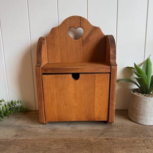 May include: A medium-brown wooden wall-mounted storage bin featuring a flat top shelf and a front-opening compartment. The back panel has a cutout heart shape at the top center, and the front door includes a small circular finger hole for opening.