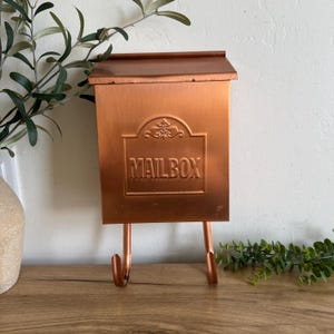 May include: A copper-colored mailbox with the word "MAILBOX" embossed on the front. The mailbox has a small roof and two hooks below. The mailbox is made of metal and has a rustic, vintage appearance. The mailbox is designed to be mounted on a wall or post.