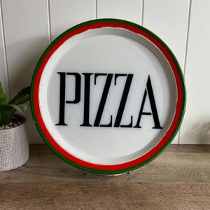 May include: A round white serving tray with a black serif font spelling the word PIZZA in the center. The tray features a thick red inner border and a dark green outer rim, creating a distinct circular frame around the central text.