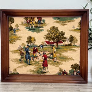 May include: A framed vintage tapestry depicting a golf scene with figures in period attire. The artwork features a brown wooden frame and a detailed landscape with trees, a house, and golfers in various poses. The color palette includes greens, browns, and reds.