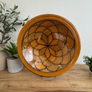 May include: A round, decorative ceramic plate with a floral design. The plate features a warm, orange-brown color with a black floral pattern in the center. The rim of the plate is a darker shade of brown. The plate is displayed on a wooden surface.