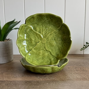 May include: A pair of round, light green ceramic plates featuring a molded cabbage leaf design with intricate veining and a glossy finish. The plates have scalloped edges that mimic the natural shape of a cabbage leaf, with one plate standing upright.