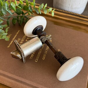 May include: A vintage door knob set featuring two smooth, round white porcelain knobs connected by a metal spindle and a silver-toned cylindrical latch mechanism with a beveled bolt. The hardware displays aged dark metal components and a classic design.