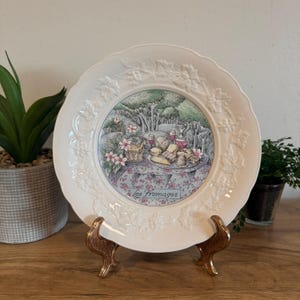 May include: A white decorative plate with a detailed illustration of a picnic scene, featuring cheese, wine, and flowers. The plate has a scalloped edge with a grape vine design and the words "Les Fromages".