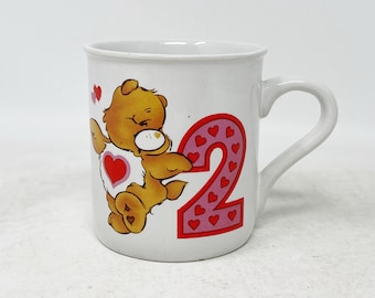 May include: White ceramic mug with a cartoon bear wearing a red heart and holding a pink number two with hearts. The mug has a handle.