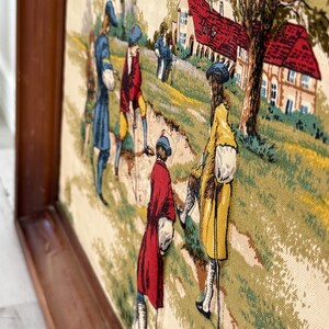 May include: A framed tapestry depicting a historical scene with figures in period clothing. The artwork features a landscape with houses and trees, and the figures are dressed in shades of blue, red, and yellow. The frame is made of dark wood.