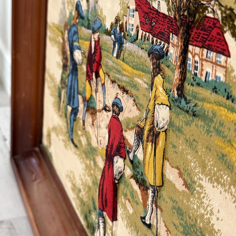 May include: A framed tapestry depicting a historical scene with figures in period clothing. The artwork features a landscape with houses and trees, and the figures are dressed in shades of blue, red, and yellow. The frame is made of dark wood.