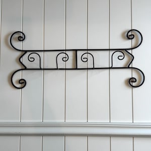 May include: Black wrought iron wall-mounted decorative rack with a scroll design. The rack has a horizontal bar with three curved hooks and decorative scrollwork on each end. The rack is mounted on a white paneled wall.