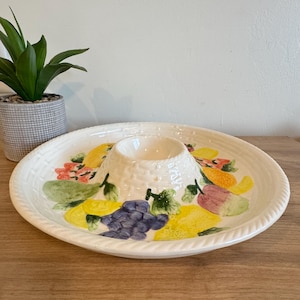 May include: A white ceramic serving platter with a central dip bowl. The platter is decorated with colorful painted fruits, including grapes, lemons, and pears. The rim has a woven texture.