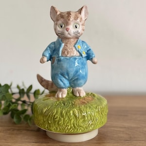 May include: A ceramic figurine of a cat wearing a blue outfit with yellow buttons, standing on a green grassy base. The cat has brown and tan fur, with a pink nose and green eyes. The figurine is a decorative item.
