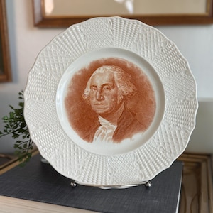 May include: A white ceramic plate with a scalloped edge and a textured basket-weave pattern on the rim. The center of the plate features a sepia-toned portrait of George Washington, depicted in a classic style with a white cravat and dark coat.