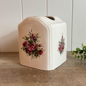 May include: A cream-colored ceramic tissue box cover with a rounded top and a beaded edge detail. The front and side panels feature a floral decal of deep pink roses with green leaves and stems, set against a smooth, glossy finish.