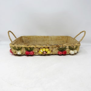 May include: A rectangular woven basket with two handles. The basket is made of natural materials and has a decorative border of flowers and berries.