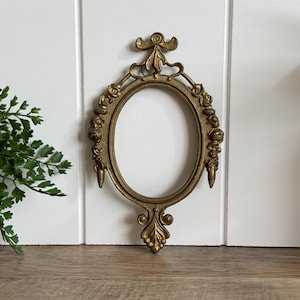 May include: An oval-shaped brass picture frame featuring intricate relief detailing along the border. The frame is adorned with sculpted floral motifs, a decorative bow at the top, and a pointed finial at the bottom, creating a symmetrical design.