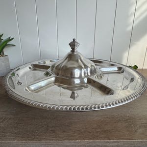 May include: A round silver-toned metal serving platter featuring a central lidded compartment with a decorative finial. The platter is divided into four distinct sections radiating from the center, with a textured rope-style border along the edge.