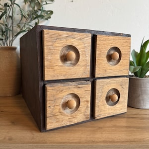 May include: A small, square wooden storage box with four drawers. The drawer fronts are light-colored wood with a circular indentation and a small wooden knob. The box's frame is a darker brown color. The box is sitting on a wooden surface.