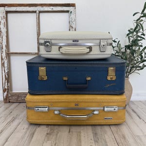 May include: Three vintage suitcases stacked on top of each other. The top suitcase is off-white, the middle one is navy blue, and the bottom one is mustard yellow. Each suitcase has a handle and metal hardware.
