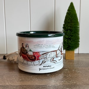 May include: A white Rival Potpourri Crock with a dark green rim. The exterior features a graphic of a horse-drawn red sleigh with two figures in a snowy landscape. A white electrical cord with a plug extends from the side of the base.