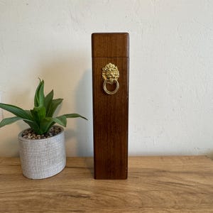 May include: A tall, rectangular wooden wine bottle holder with a brass lion's head and ring handle. The holder is a rich brown color and stands upright on a wooden surface. A small potted plant is to the left of the holder.