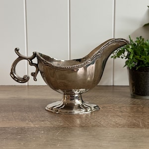 May include: A silver-toned gravy boat featuring a polished metallic finish, a curved handle with ornate detailing, a wide pouring spout, and a pedestal base. The rim of the vessel is decorated with a beaded texture, and the body has a smooth, reflective surface.