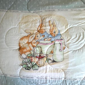May include: A quilted blanket featuring a charming illustration of three rabbits gathered around a basket of berries and a pitcher. The design includes the text "Once upon a time" in a repeating pattern. The color palette includes soft pastels.