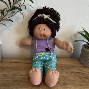 May include: A vintage doll with brown hair tied with a white ribbon. The doll wears a purple top with a gray vest and blue pants with white and green polka dots. The doll is sitting on a wooden surface.