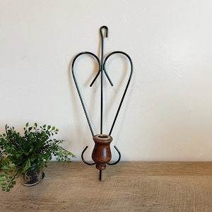 May include: Black metal wall sconce with a wooden candle holder. The sconce has a heart-shaped design and a hook at the top.