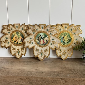 May include: Three decorative wall plaques shaped like large leaves with gold and cream textured finishes. Each plaque features a central circular print depicting 18th-century figures in outdoor settings, accented with small green diamond-shaped inlays.