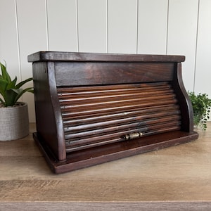 May include: A dark brown wooden bread box featuring a roll-top tambour door with horizontal slats. The rectangular storage container has a flat top surface, a solid base, and a small metallic handle centered on the front of the sliding door.