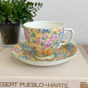May include: A floral teacup and saucer set. The cup and saucer feature a yellow background with pink, purple, blue, and orange floral patterns. The rim and handle of the cup are outlined in green.