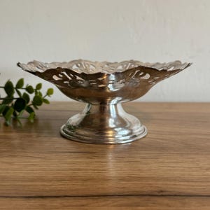 May include: A silver-colored, pedestal-style serving dish. The dish has a scalloped edge with decorative cut-out patterns. The base is a short, round pedestal. The dish is likely made of metal and is suitable for displaying small items.