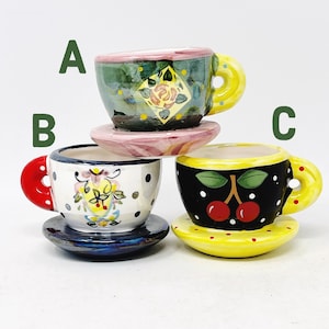 May include: Three decorative ceramic teacups with matching saucers. Each cup features a unique design: a green cup with a floral pattern, a white cup with polka dots and a floral design, and a black cup with cherries. The handles and saucers are in various colors.