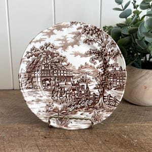 May include: A round ceramic plate featuring a brown and white transferware pattern of a horse-drawn carriage in front of a timber-framed building. The scene includes trees, a wooden fence, and a stream, with a scalloped edge design.