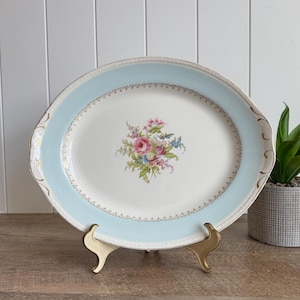May include: An oval ceramic serving platter featuring a pale blue border with gold filigree detailing. The center of the white platter displays a colorful floral bouquet with pink roses, blue forget-me-nots, and green foliage accents.