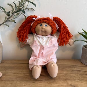 May include: A vintage doll with a soft, fabric body and a vinyl face. The doll has bright orange yarn pigtails tied with white ribbons, blue eyes, and a pink dress with white lace trim. The doll is sitting on a wooden surface.