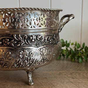 May include: A silver-toned metal bowl featuring an intricate embossed floral pattern across its body, a decorative openwork lattice rim with a twisted rope edge, two side handles, and three small feet supporting the base of the vessel.