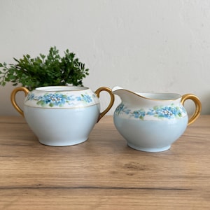 May include: A vintage light blue and white porcelain sugar bowl and creamer set. The set features gold handles and rims, with a floral design of blue flowers. The sugar bowl has a lid, and the creamer has a spout.