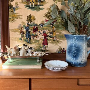 May include: A wooden framed print with a pastoral scene, a silver dog figurine tray, a small decorative dish, and a blue pitcher with foliage. The dog figurines are in various poses. The pitcher is blue with a floral design.