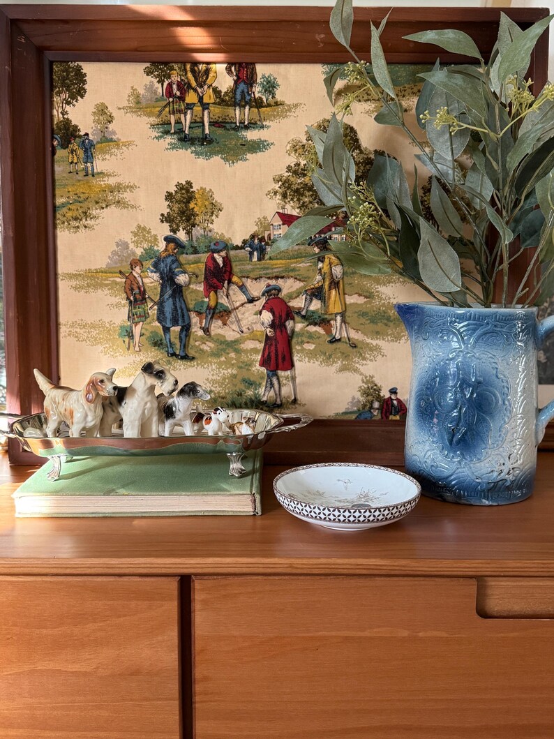 May include: A wooden framed print with a pastoral scene, a silver dog figurine tray, a small decorative dish, and a blue pitcher with foliage. The dog figurines are in various poses. The pitcher is blue with a floral design.