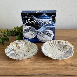 May include: Silverplated shell trays, two displayed in front of a box. The trays are shaped like nautilus shells with detailed ridges and a shiny silver finish. The box reads "Silverplated Shell Trays."