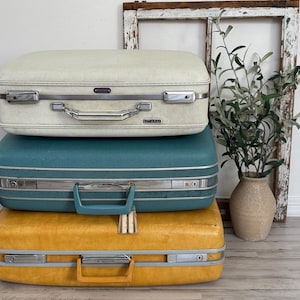May include: Three vintage Samsonite suitcases stacked on top of each other. The top suitcase is off-white, the middle one is teal, and the bottom one is yellow. Each suitcase has a silver handle and trim.